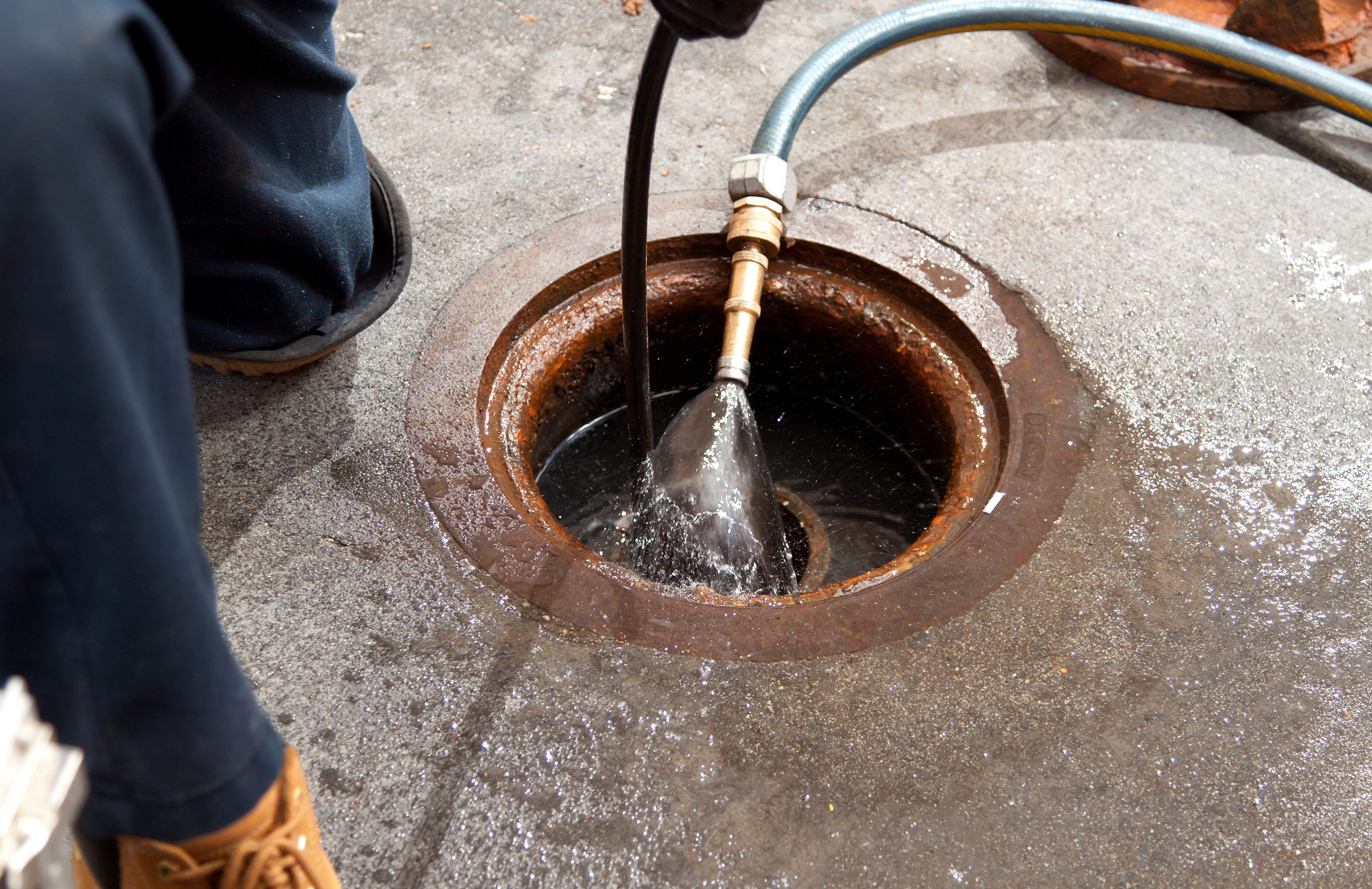 Worker Kneeling Next To Sewer Opening To Find Clog