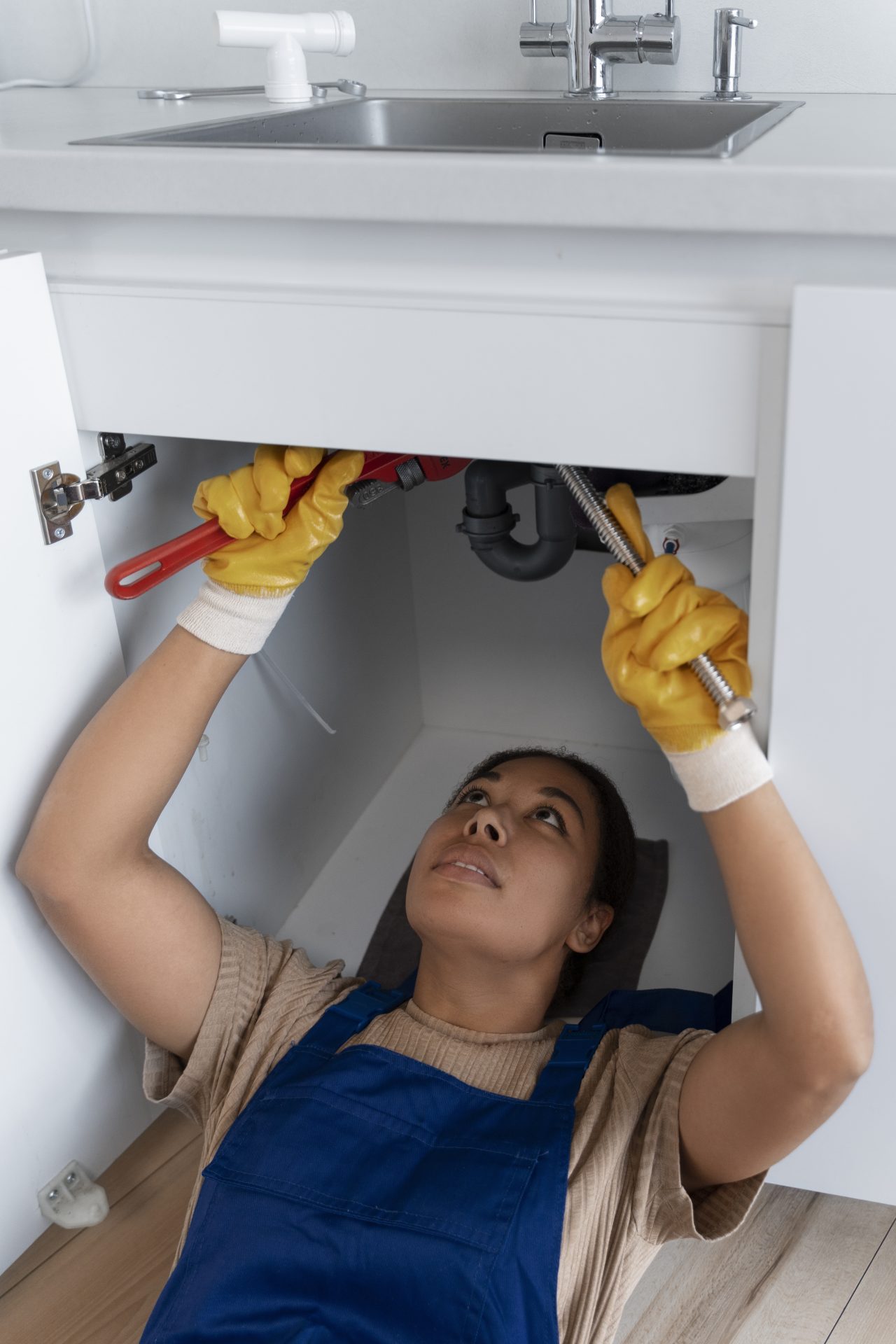 A female plumber is lying on the floor underneath a sink, looking up with high concentration at the drain and fixing it with a tool