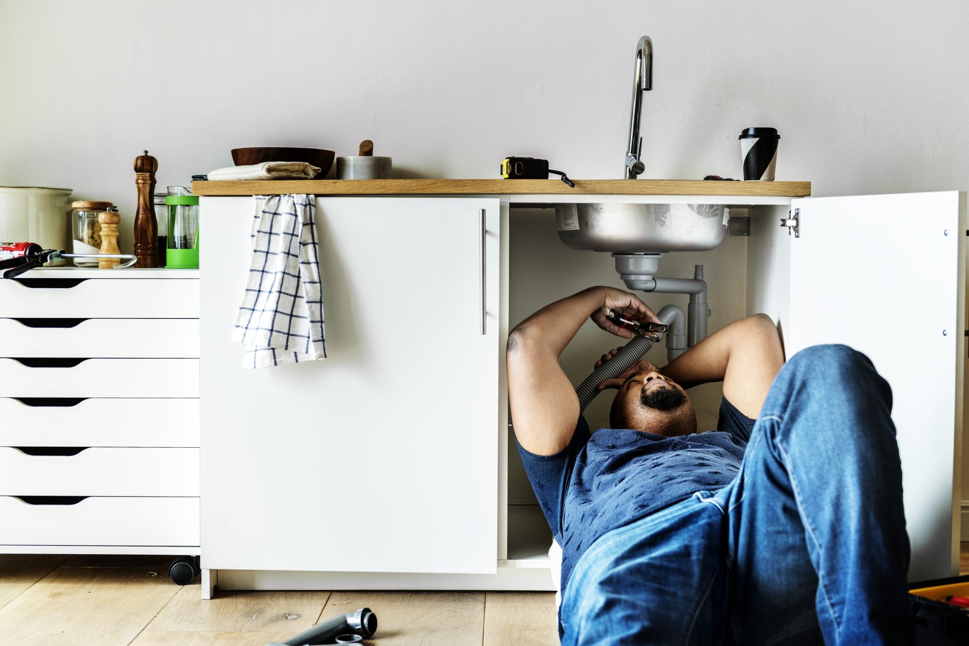 A male Plumber wearing jeans is lying down underneath a kitchen sink and working on the pipe.