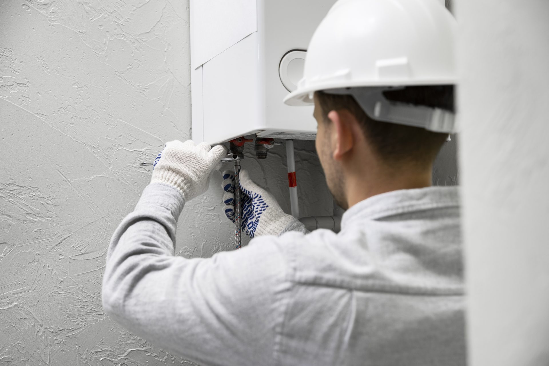 A male technician wearing a white helmet faces a modern, white water heater suspended from a wall and is fixing it.