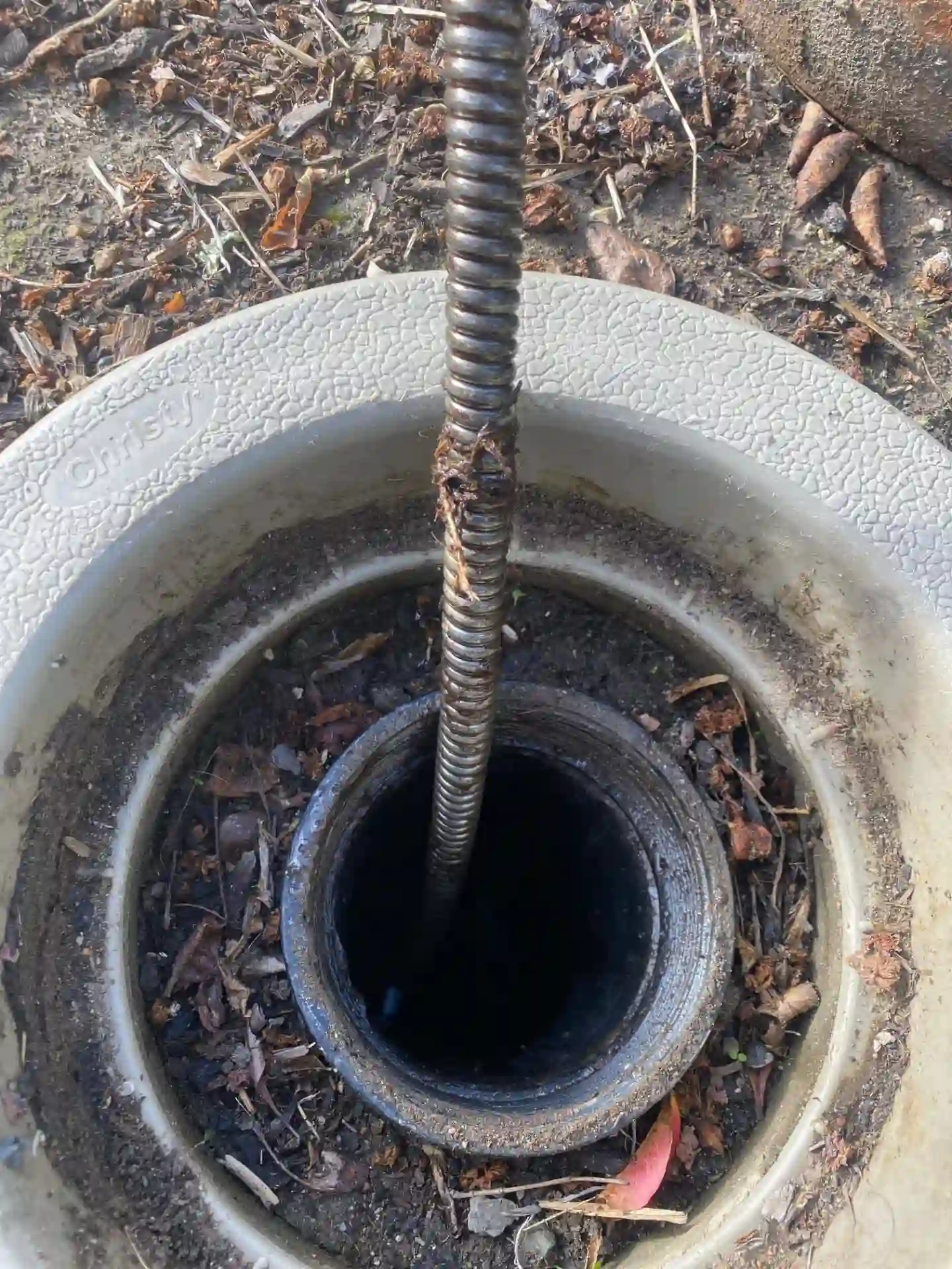 A drain auger is clearing a blockage through an outdoor sewer cleanout.