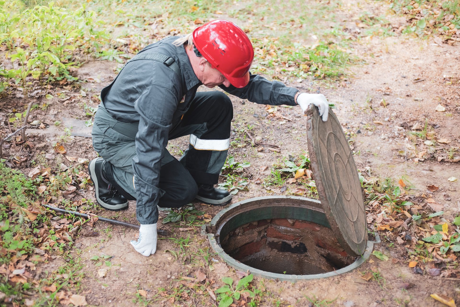 An adult man in a red hard hat kneeling on the ground and opening a septic tank hatch and peering inside