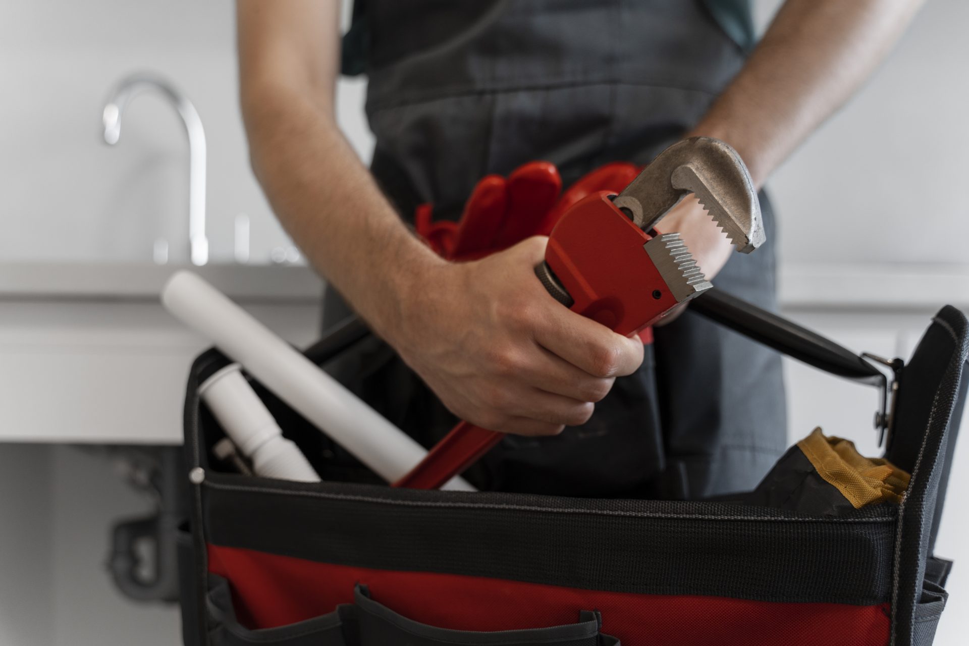 Close-up view of a man’s hand holding a red pipe wrench with a toolbag filled with plumber’s tools in the background