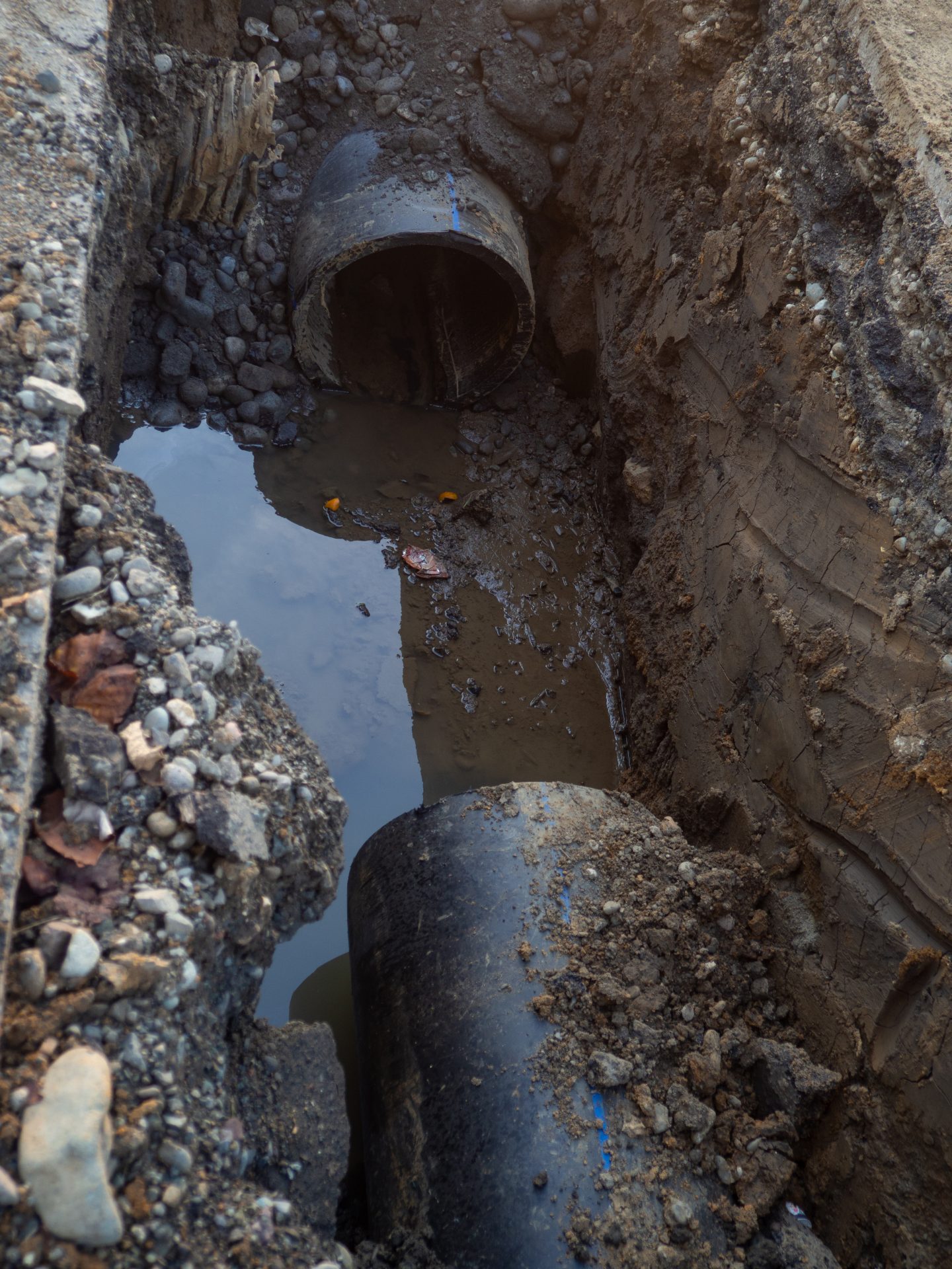 A black pipe has been partially dug up, and a large section of it is missing. There is a puddle of water between the open ends of the pipe, and it is surrounded by mud, dirt, and small stones.
