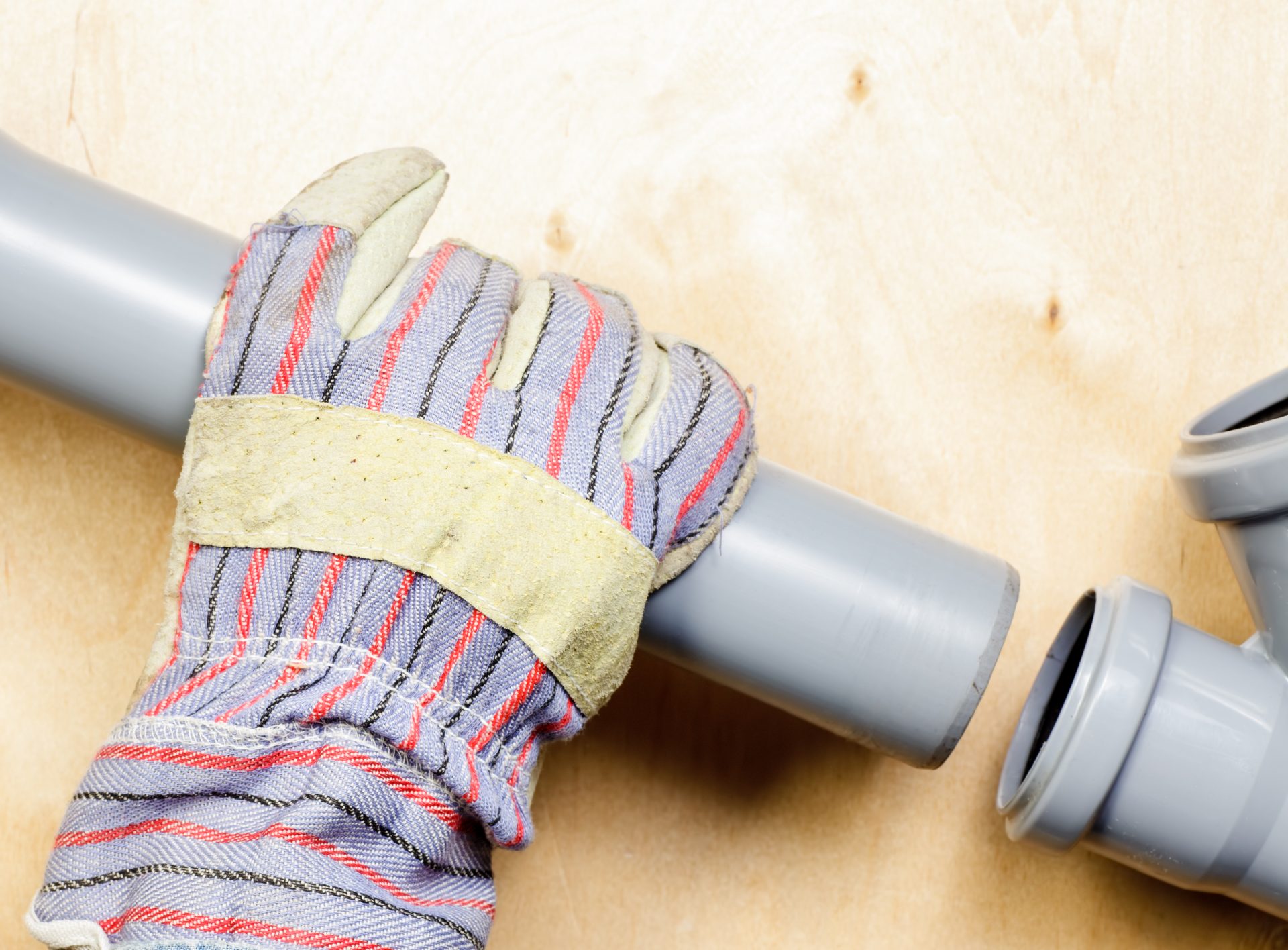 A person wearing multi-colored work gloves is holding a section of light gray piping against a tan colored wooden surface. A second Y-shaped piece of pipe is partially visible on the right side of the image.