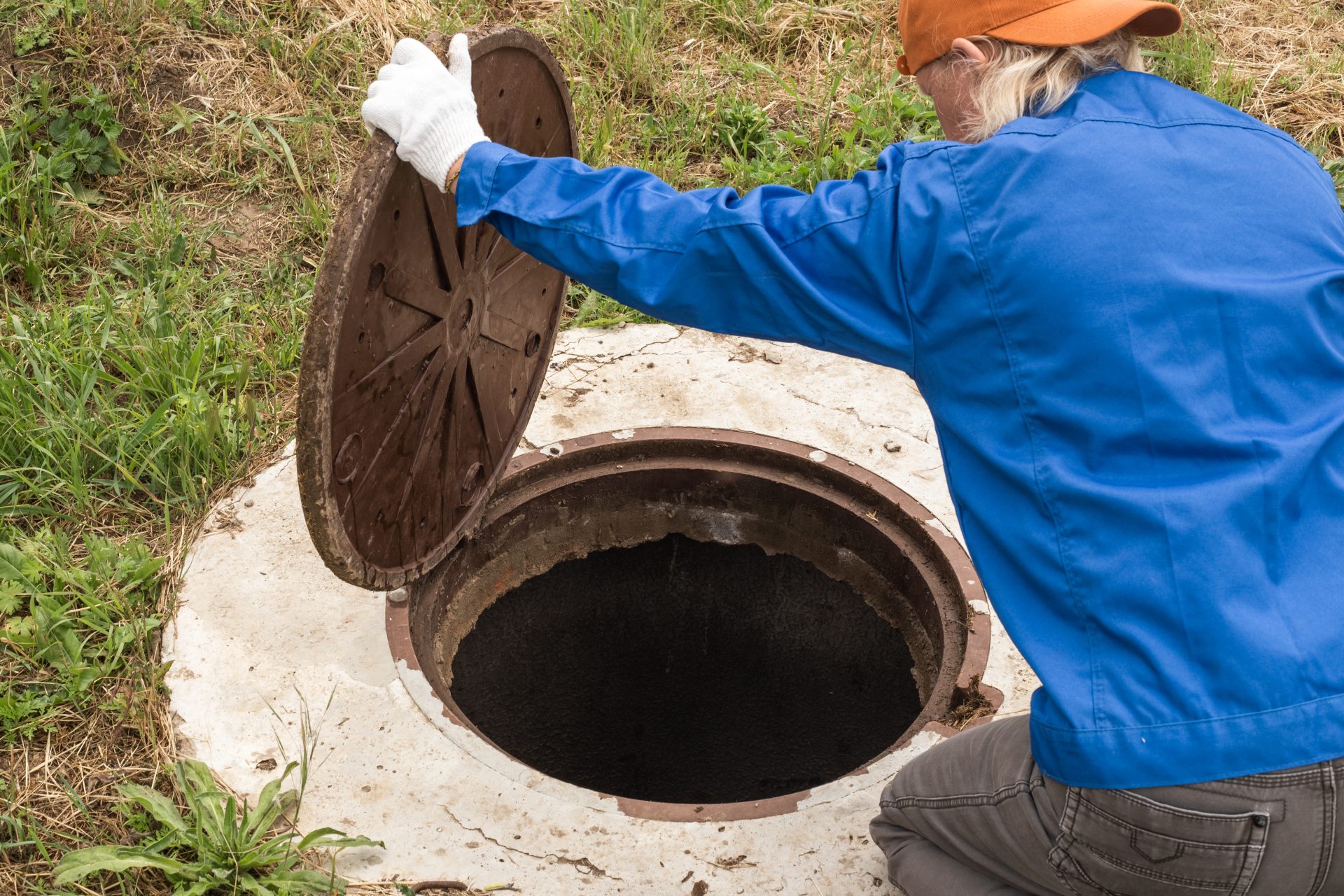 A blonde man in a blue jacket and orange hat is kneeling next to the opening above a septic tank. He's holding the circular lid open and looking inside the hole.
