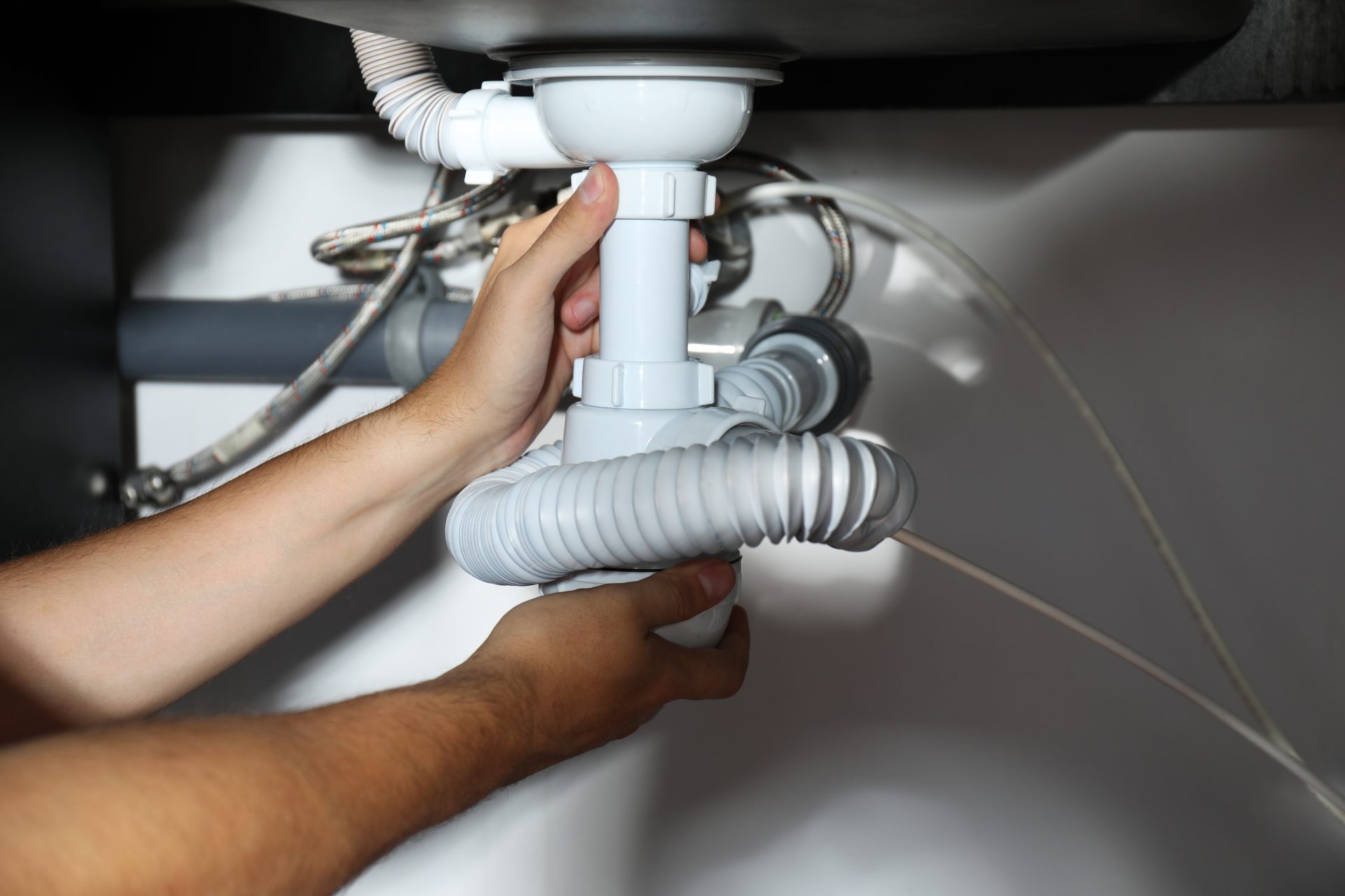 A close-up of someone's hands adjusting plastic piping beneath a sink. There are multiple pipes and accordion tubes connected to promote the proper flow of water.