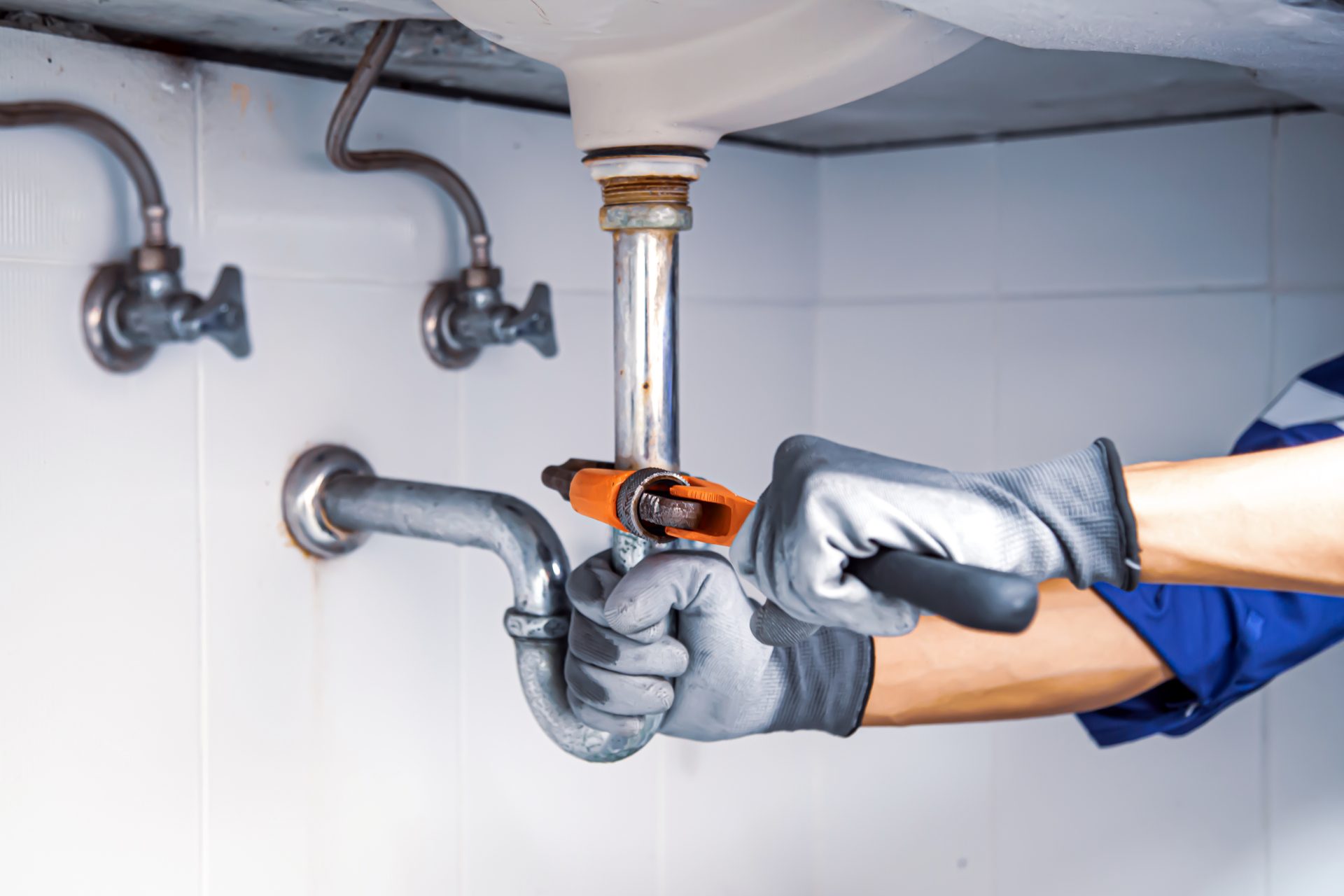 A man wearing gray work gloves and a blue long-sleeved shirt uses an orange wrench to work on a rusty silver metal pipe beneath a sink.