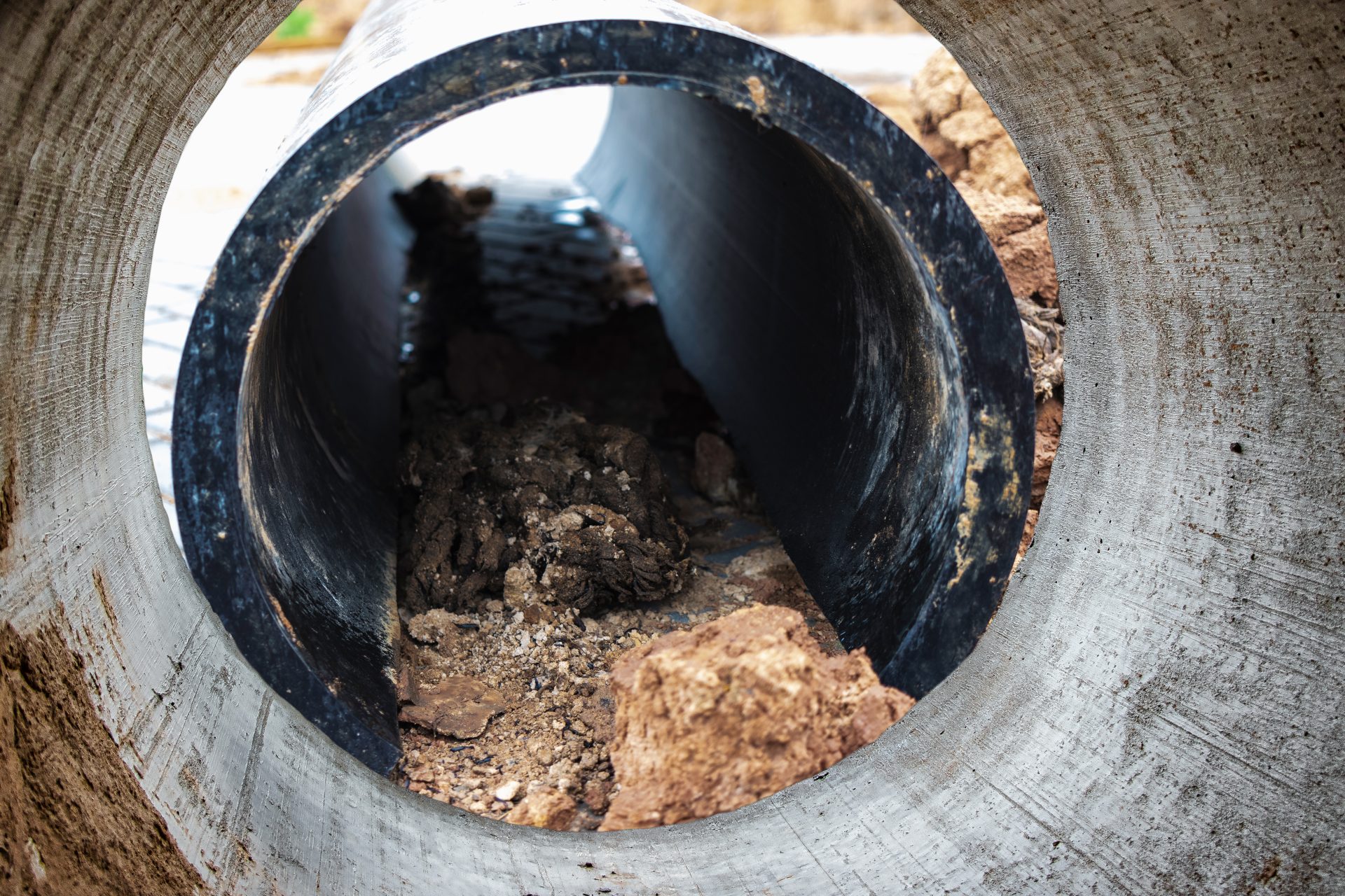 A close-up of someone's hands using a wrench to tighten a connection area on a silver, metal pipe beneath a sink. The pipe's U-shaped bend is visible beneath the place being worked on.