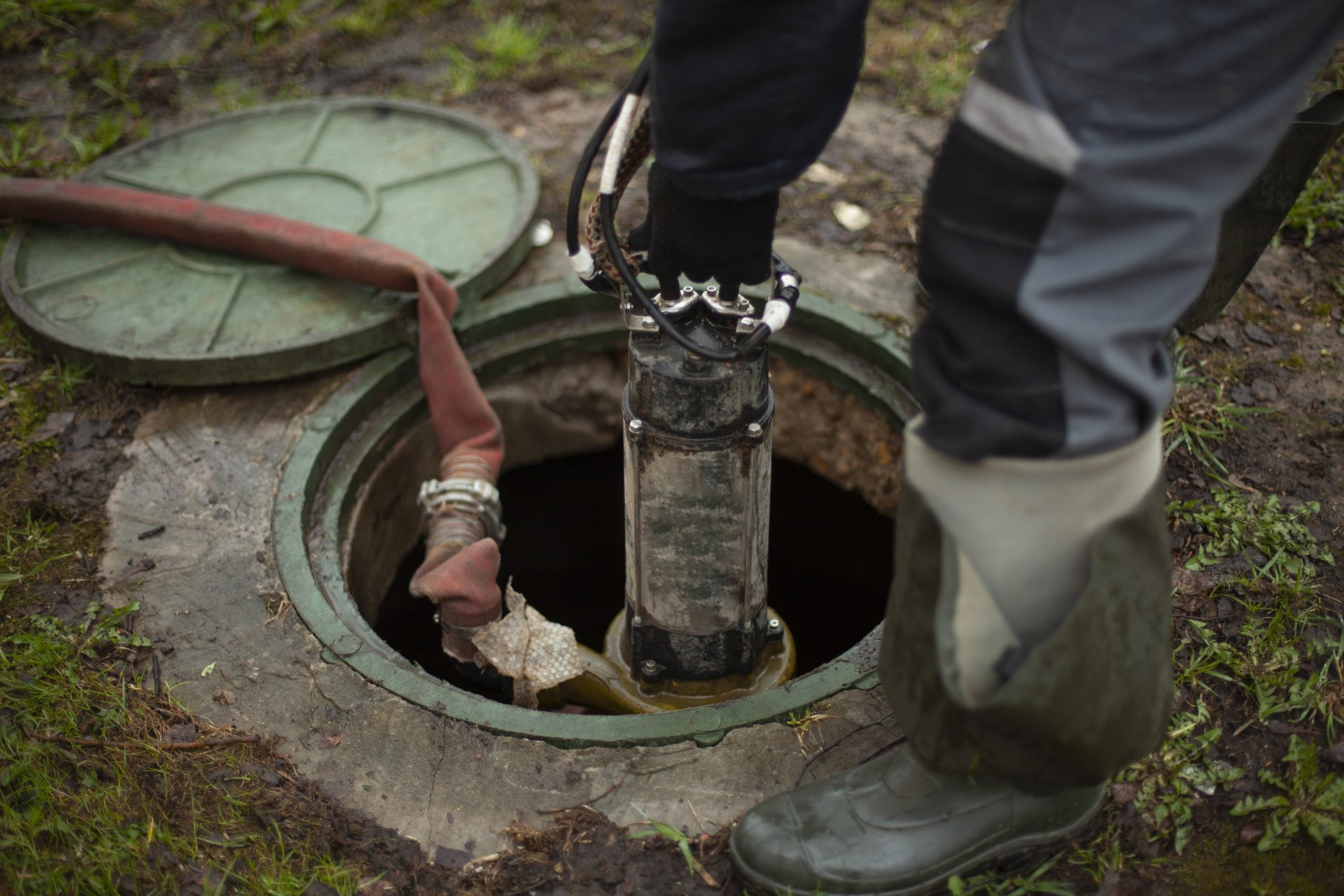 A person is using a pump inside an open septic tank access port. The hose is red, and the lid of the tank is green. The person is wearing high boots and a black and blue protective suit.