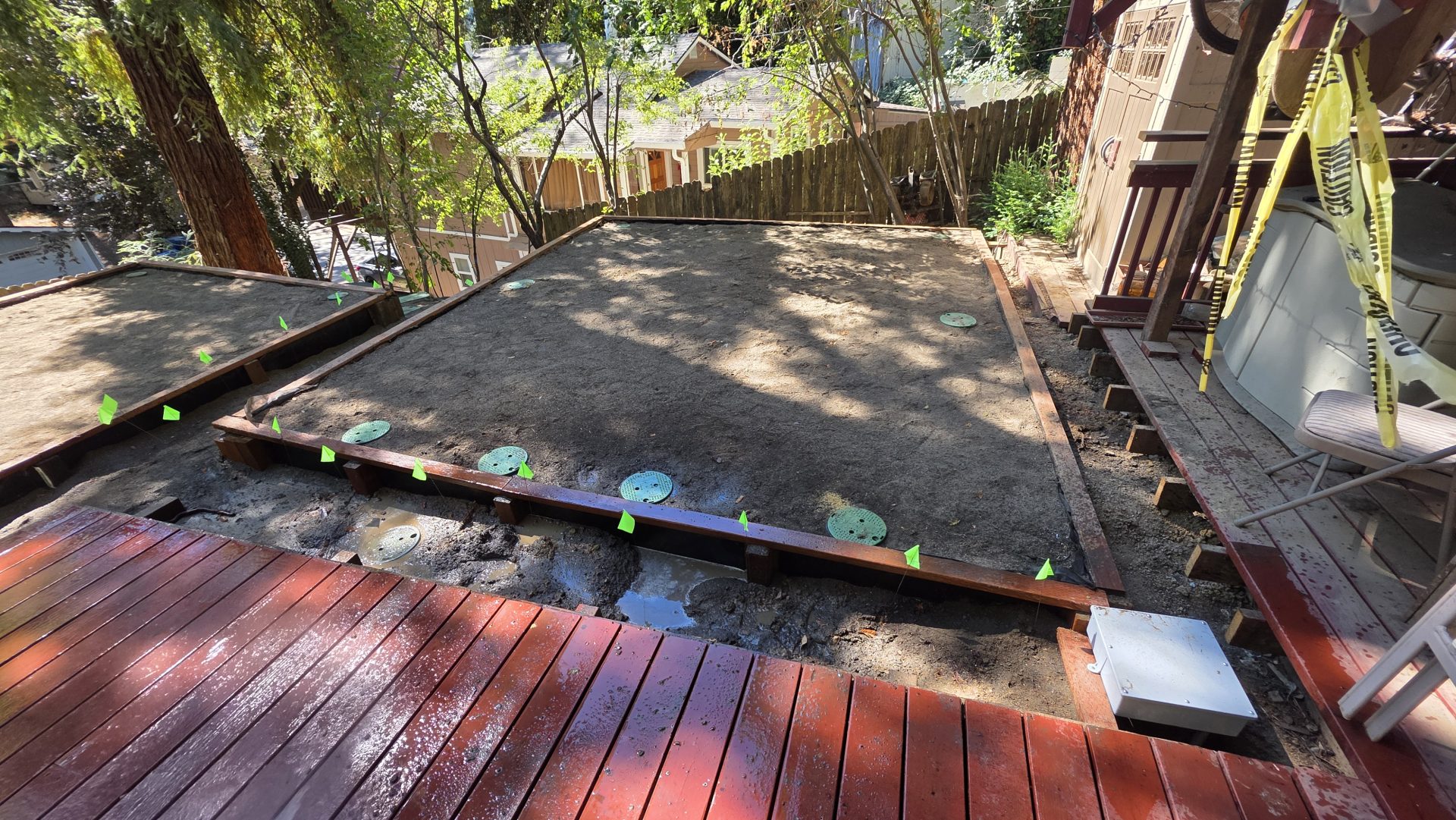 An outdoor excavation area features green access covers set in soil alongside wooden decking for specialized plumbing & rooter services.