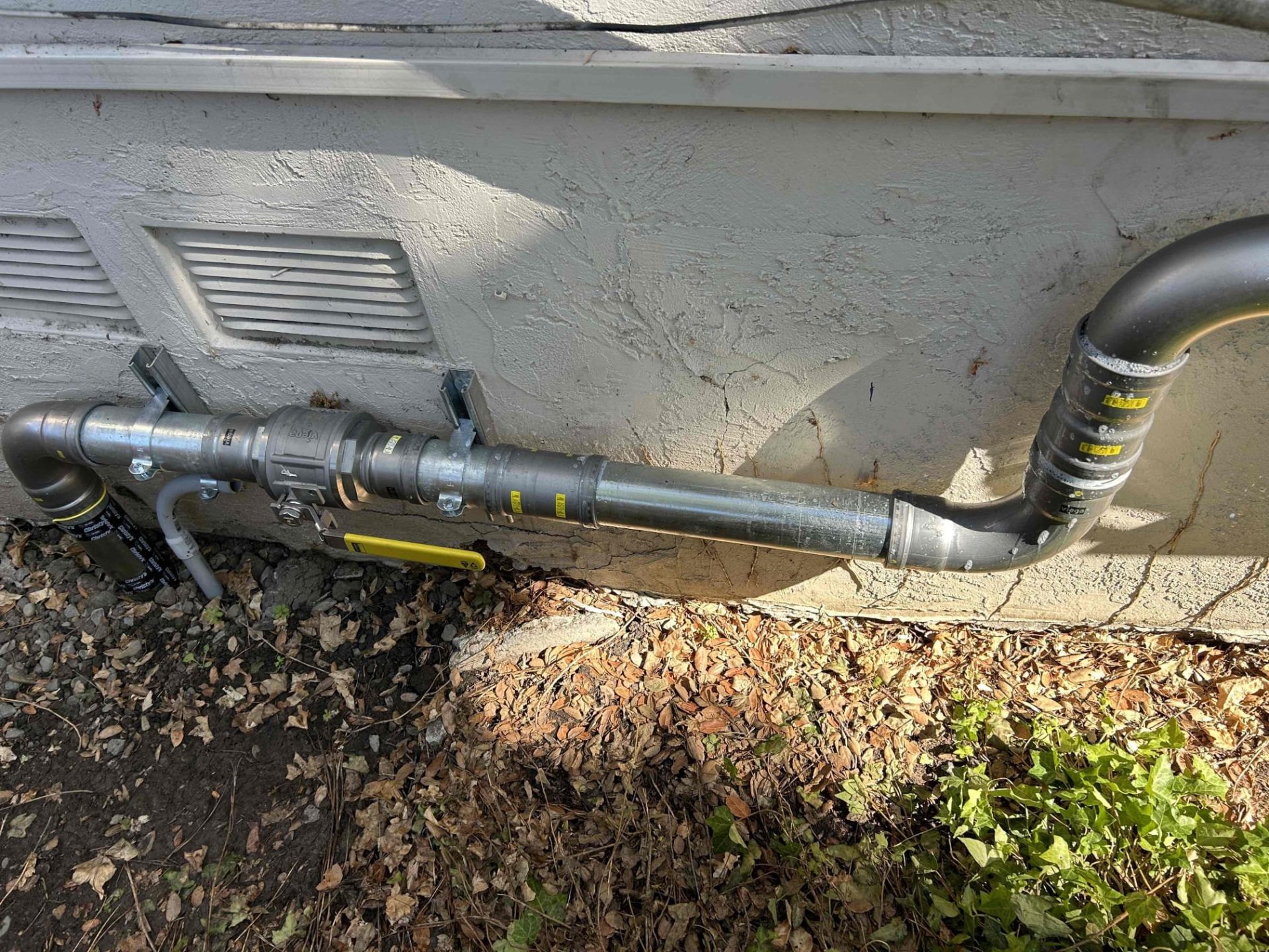 A newly installed exterior pipe assembly with a yellow handle valve runs along a building foundation for expert plumbing & rooter services.